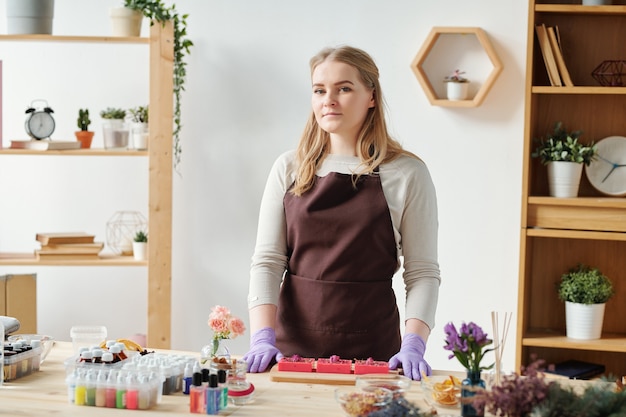 Hübsches Mädchen in Schürze und Handschuhen, die Sie beim Stehen am Tisch während der Seifenherstellung Meisterklasse betrachten