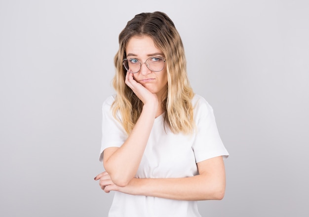 hübsches Mädchen in schwarzer Brille, das ihr Gesicht berührt und sich bückt, unter ihren Augen hervorschaut, es hasst, mit einer langweiligen Person zu sprechen und an einer grauen Wand zu stehen