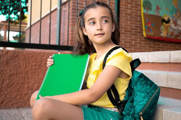 Hübsches Mädchen mit hellen Augen umklammert ein grünes Notizbuch und sitzt beim Klo auf der Schultreppe