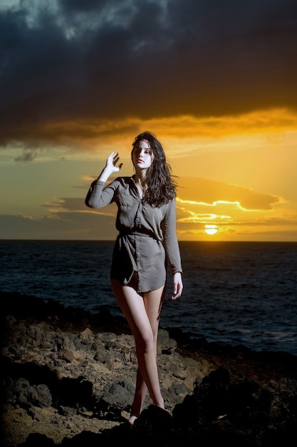 Hübsches Mädchen oder schöne Frau mit langen brünetten Haaren, die ein sexy braunes Hemd tragen, das am felsigen Ufer am blauen Meer oder Ozean während des idyllischen Sonnenuntergangs unter dunklem bewölktem Himmel auf hellgelbem Himmelshintergrund steht standing