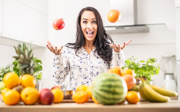 Hübsches Mädchen wirft zu Hause Apfel und Orange mit einem Küchentisch voller verschiedener Obstsorten.