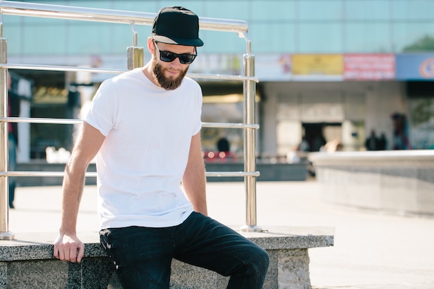 Hübsches männliches Hipster-Modell mit Bart, das weißes leeres T-Shirt und eine Baseballkappe mit Platz für Ihr Logo oder Design im lässigen urbanen Stil trägt