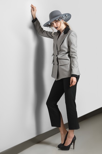 Hübsches Mode-Modell in der Jacke, in den Hosen und im Breitkrempenhut mit den Blumen, die nahe grauer Wand aufwerfen