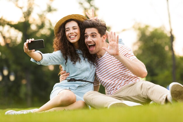 hübsches Paar, das auf grünem Gras im Park sitzt und Selfie auf Smartphone nimmt