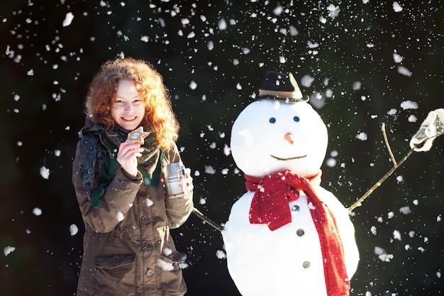 Foto hübsches rothaariges mädchen, das tee trinkt und lebkuchen im freien mit einem schneemann isst, während es schneit