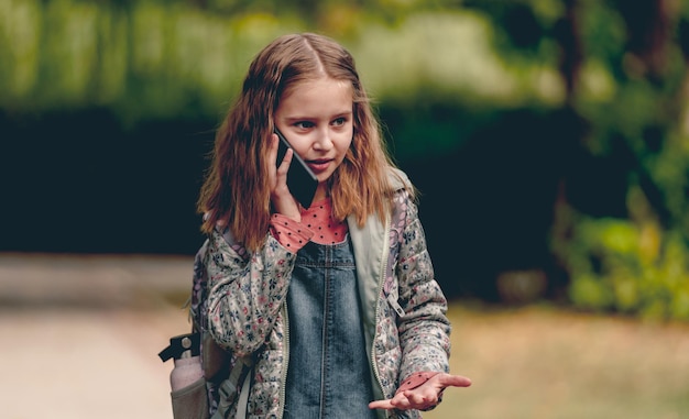 Hübsches Schulmädchen, das draußen mit dem Smartphone spricht und lächelt. Nettes weibliches Kind kommuniziert mit Handy im Herbstpark