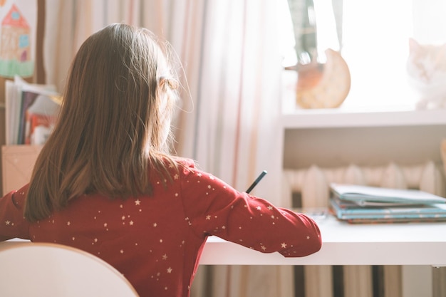 Hübsches Schulmädchen mit langen Haaren im roten Kleid zeichnet mit Bleistift am Tisch im Kinderzimmer zu Hause