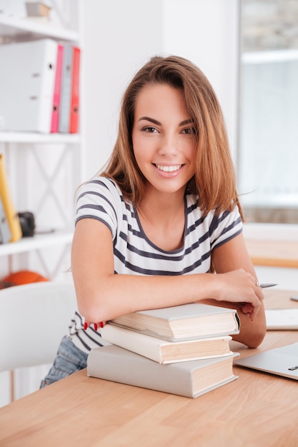 Hübsches Studentenmädchen, das in einem T-Shirt mit Streifenmuster gekleidet ist, hat die Ellbogen auf Bücher gelehnt, während es nach vorne schaut und lächelt