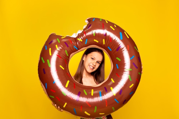 Hübsches Teenager-Mädchen blickt durch einen aufblasbaren Donut-Ring vor gelbem Hintergrund in die Kamera