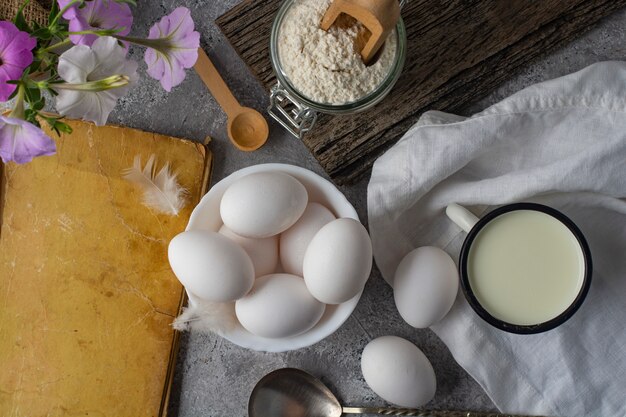 Hühnereier, Mehl, Milch und ein altes Rezeptbuch auf dem Küchentisch