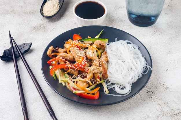 Hühnerfleisch mit Gemüse-Wok und chinesischen Reisnudeln, Saucen und Sesam in einer schwarzen Schüssel mit