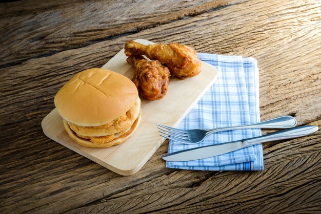 Hühnerhamburger und gebratenes Huhn auf hölzernem Schneidebrett mit Messer und Gabel, Serviette