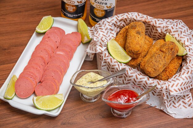 Hühnerkrokette und Cambui-Wurst, typischer brasilianischer Snack, serviert mit Zitronenscheiben, Chilisauce und Mayonnaise auf Holztisch, serviert mit berühmtem Corona-Bier