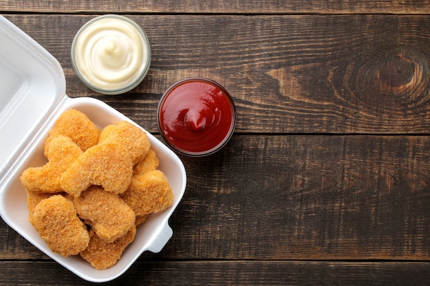 Hühnernuggets in einer Lebensmittellieferungsbox mit weißer und roter Soße auf braunem Holzhintergrund. Fast-Food-Draufsicht mit Platz für Text