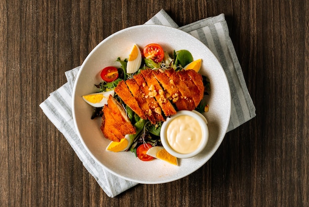 Hühnerschnitzel, serviert mit frischem Salat, gekochten Eiern, Tomaten und Mayonnaise in weißer Schüssel auf dunklem Holz