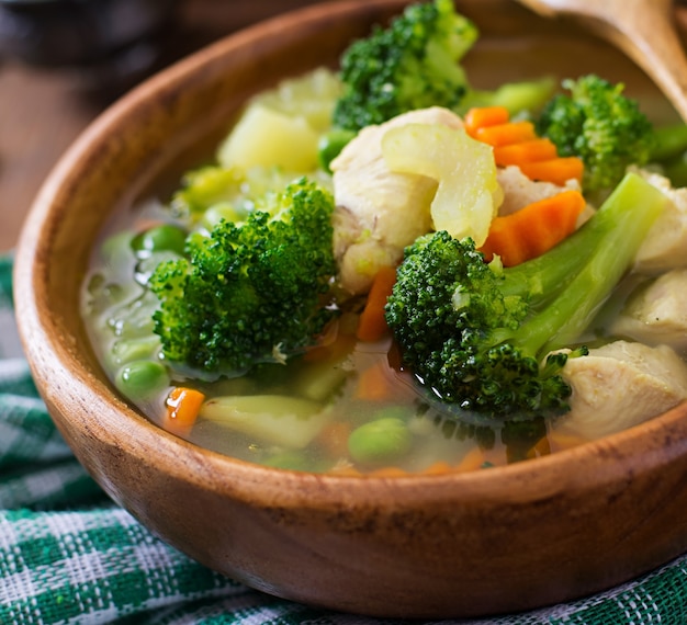 Hühnersuppe mit Brokkoli, Erbsen, Karotten und Sellerie in einer Schüssel auf einer Holzoberfläche im rustikalen Stil