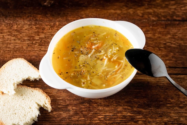 Hühnersuppe mit Nudeln, Kartoffeln, Zwiebeln und Karotten in einem weißen tiefen Teller mit schwarzem Löffel und Weißbrot auf Holzhintergrund