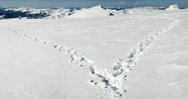 La huella humana forma la forma del corazón en la meseta de la ladera cubierta de nieve y las cadenas montañosas detrás.