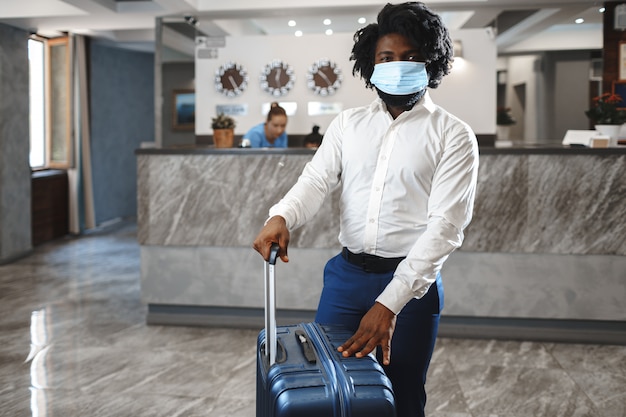Huésped del hotel hombre africano con maleta con máscara protectora para protegerse del coronavirus