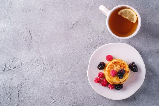 Hüttenkäse Pfannkuchen Quark Krapfen Dessert mit Himbeere und Brombeere Beeren in Platte in der Nähe von heißen Teetasse mit Zitronenscheibe auf Stein Beton Hintergrund Draufsicht Kopie Raum