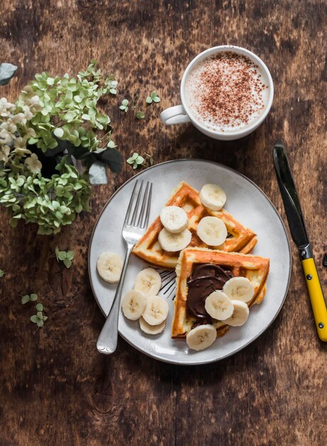 Hüttenkäse Waffeln mit Schokoladenpaste Banane und Cappuccino auf einem hölzernen Hintergrund Draufsicht Köstliches Frühstücksdessert