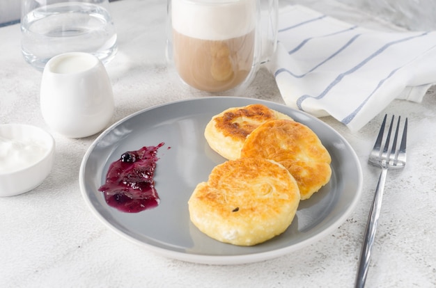 Hüttenkäsepfannkuchen auf einem schwarzen Teller mit Sahne, Marmelade und Tasse Latte. Gesundes Frühstück. Hausgemachtes Essen