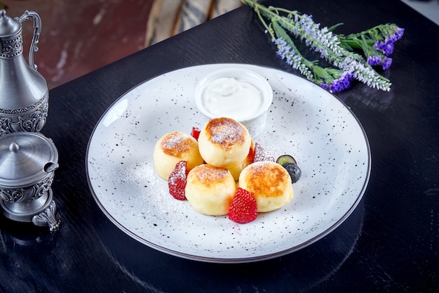 Hüttenkäsepfannkuchen serviert auf weißem Teller mit heißer saurer Sahne und Erdbeere auf dunklem hölzernem Hintergrund. Traditionelles hausgemachtes ukrainisches Essen Syrniki. Gourmet-Frühstück. Dessertküche