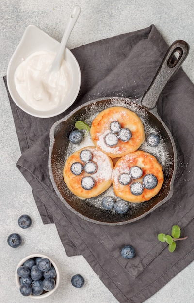 Hüttenkäsepfannkuchen, Syrniki, Quarkstückchen mit frischen Heidelbeeren, Puderzucker und Sauerrahm in einer gusseisernen Pfanne