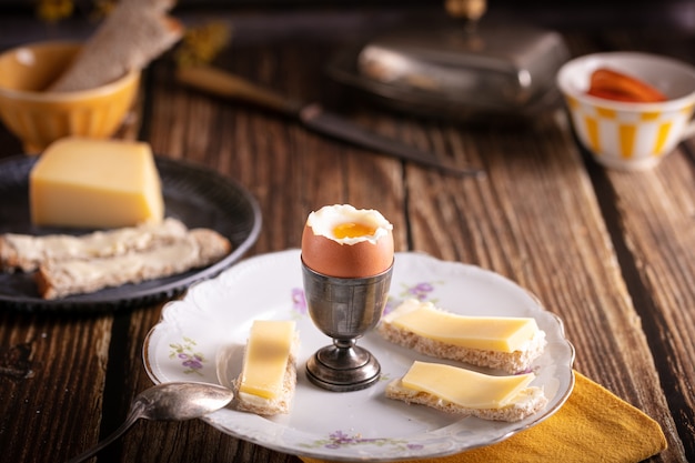 Huevo cocido en una huevera plateada con pan y queso sobre una mesa de madera