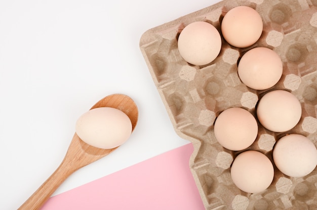 Huevo en una cuchara de madera. Una bandeja de huevos sobre un fondo blanco y rosa. bandeja ecológica con testículos. Tendencia minimalista, vista superior. Bandeja de huevos. Concepto de pascua.