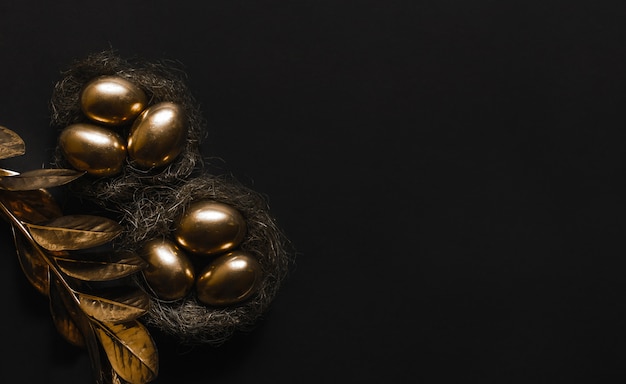 El huevo está pintado de oro en un nido de paja y la planta con hojas doradas sobre una mesa negra.