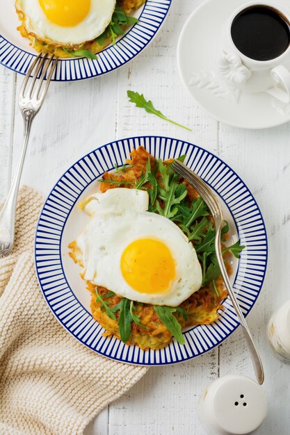 Huevo frito con panqueque de papa, rúcula y aguacate en un plato de cerámica para el desayuno en la superficie de la mesa de madera blanca