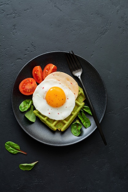 Huevo frito con queso, tomates, acelgas y gofres con espinacas en placa de cerámica negra sobre mesa de hormigón oscuro. Sándwich para el desayuno. Enfoque selectivo. Vista superior. Copie el espacio.