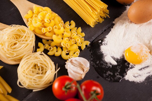 Huevo en harina junto a diferentes tipos de pasta cruda sobre fondo oscuro