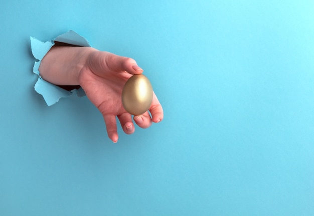 Foto huevo de oro en una mano femenina sobre un fondo azul con un agujero, concepto de beneficio