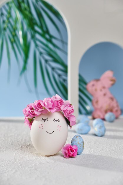 Huevo de pascua blanco natural con cara dulce dibujada en la playa tropical con muchos pequeños arcos de huevos azules y una palmera en una luz suave Tarjeta de vacaciones de Pascua feliz