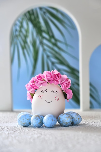 Huevo de pascua blanco natural con cara dulce dibujada en la playa tropical con muchos pequeños arcos de huevos azules y una palmera en una luz suave Tarjeta de vacaciones de Pascua feliz