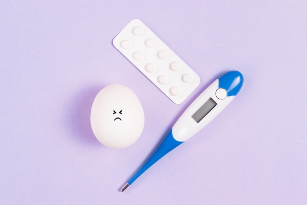 Huevo de Pascua con una cara triste con un termómetro y un blister de pastillas sobre un fondo azul.