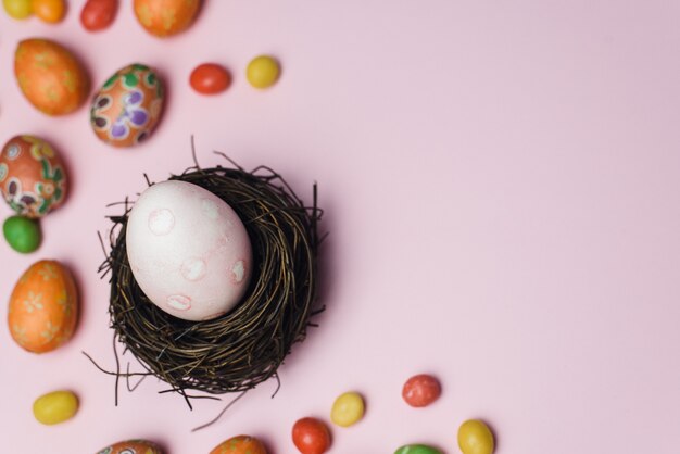 Huevo pintado para las vacaciones de Semana Santa en un nido de ramas junto a dulces y galletas de jengibre. Tarjeta de Pascua sobre un fondo rosa