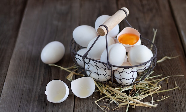 Huevos blancos orgánicos frescos con cáscara de huevo y yema en canasta de malla metálica sobre fondo de madera y paja