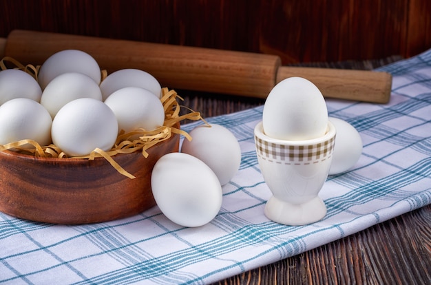 Huevos blancos en un tazón de madera y sobre una toalla rústica sobre una mesa de madera. Hay un rodillo en el fondo. Clave baja.