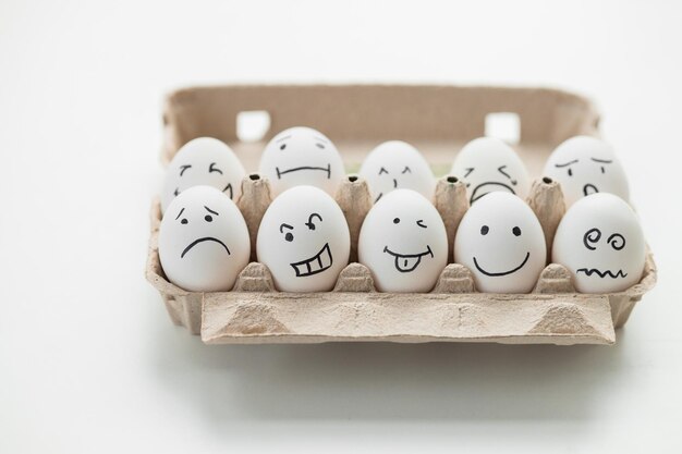 Huevos con caras graciosas en envase de cartón sobre mesa de madera blanca