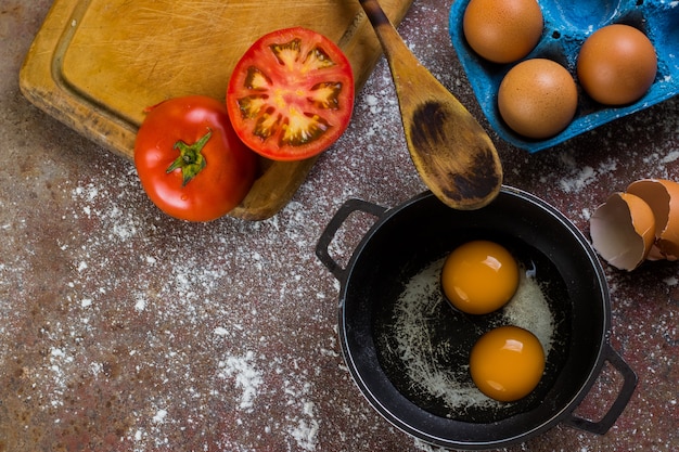 huevos crudos en sartén o olla acompañados de tomate y huevos frescos en tabla de cortar y cuchara de madera