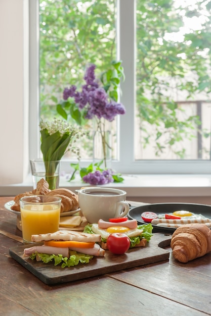 Huevos fritos con salchicha y verduras en sartén. Desayuno con zumo de naranja y croissant sobre tablas rústicas de madera, superficie de alimentos.