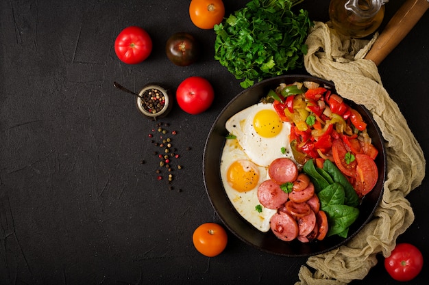 Huevos fritos con salchichas y verduras en una sartén sobre una mesa negra en estilo rústico.