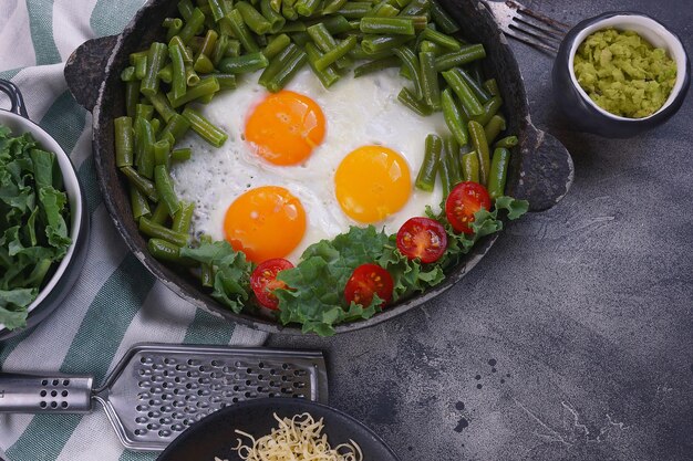 Huevos fritos en una sartén vieja con tomates, pesto de aguacate, judías verdes, pimienta y ensalada sobre un fondo oscuro