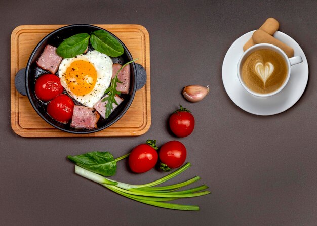 Huevos fritos con tomate y jamón en una sartén sobre una tabla y una taza de capuchino con corazón