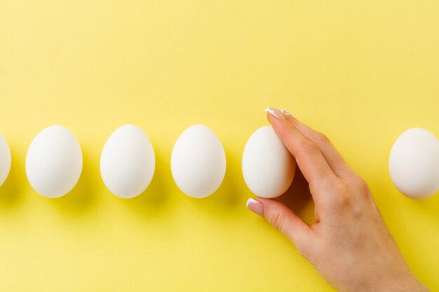 Huevos de gallina cruda blanca sobre superficie de luz amarilla y mano femenina sostiene huevo roto. Vista superior.