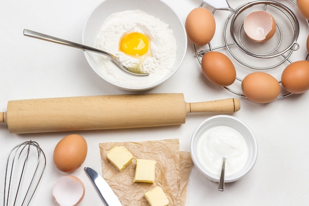 Huevos de gallina marrón en un recipiente de cartón, harina, huevo roto y cuchara en un tazón sobre fondo blanco.