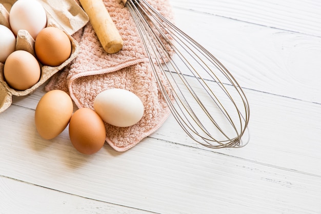 Huevos de gallina orgánicos en cajas de papel marrón, batidor y rodillo colocado sobre una mesa de madera blanca. Materias primas y equipos para hacer pan casero Vista superior y espacio de copia